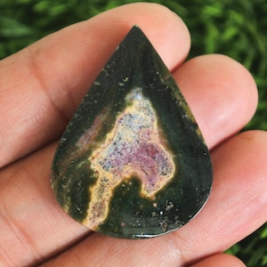 Pode incluir: Uma pedra preciosa em forma de gota, com um exterior verde escuro e um padrão central de roxo, amarelo e branco. A pedra polida é segurada na mão, mostrando suas cores naturais e design único. Ideal para fazer joias.