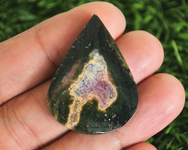 May include: A teardrop-shaped gemstone with a dark green exterior and a central pattern of purple, yellow, and white. The polished stone is held in a hand, showcasing its natural colors and unique design. This gemstone is ideal for jewelry making.