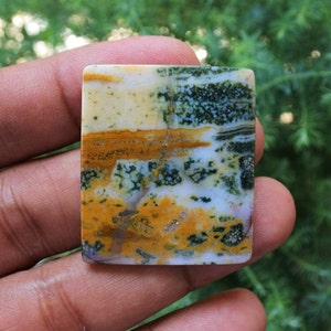 May include: A square gemstone displaying a natural pattern of colours. The stone has a mix of yellow, orange, green, and white, resembling a landscape. The polished surface of the gemstone is visible, held in a hand, highlighting its natural beauty.