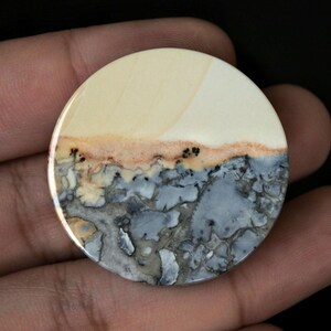 May include: A round, polished stone with a unique pattern. The top half is a creamy white, transitioning to a band of tan and then a lower section of gray and blue mottled stone. Small black specks are scattered throughout the tan band.