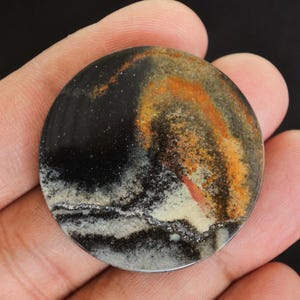 May include: A circular, polished stone featuring a dark, speckled backdrop and swirling patterns of orange, tan, and white. The stone has a metallic silver edge and is held in a hand.