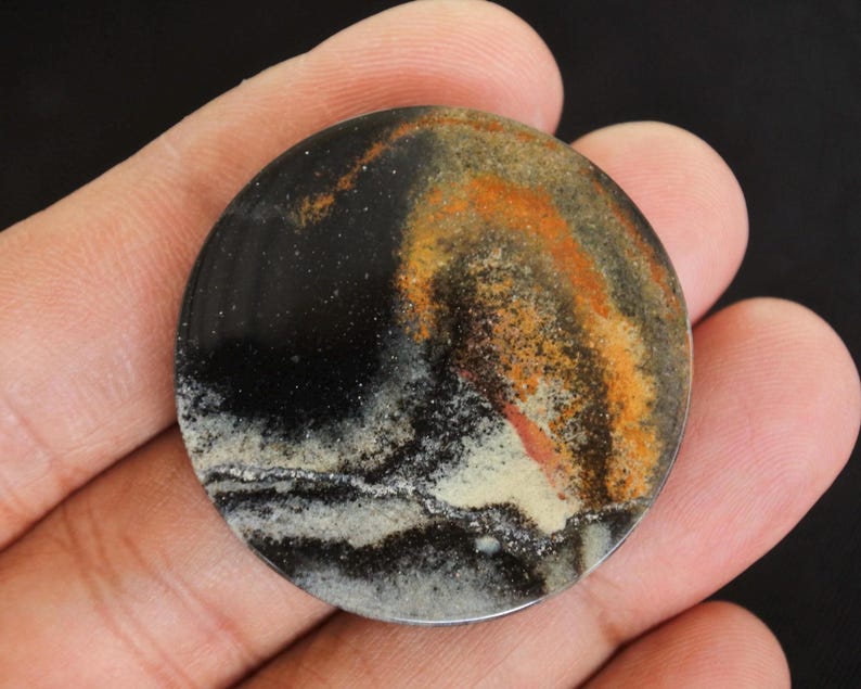 K&ouml;nnte beinhalten: Ein runder, polierter Stein mit einem dunklen, gesprenkelten Hintergrund und wirbelnden Mustern in Orange, Beige und Wei&szlig;. Der Stein hat einen metallischen Silberrand und wird in einer Hand gehalten.