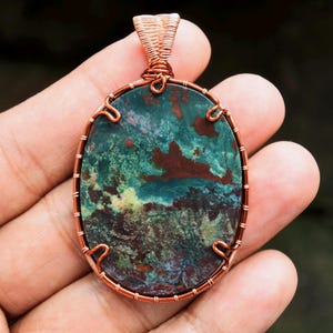 Peut inclure: Pendentif ovale avec un cadre en fil de cuivre. Le pendentif présente une pierre aux couleurs mélangées de turquoise, de vert et de marron. Le fil de cuivre est tissé de manière complexe autour de la pierre et forme une boucle pour une chaîne.