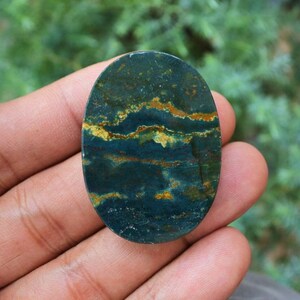 K&ouml;nnte beinhalten: Ein ovaler Edelstein mit einer dunkelblauen Grundfarbe, der goldene und braune B&auml;nder aufweist. Der polierte Stein hat ein nat&uuml;rliches, erdiges Aussehen. Der Edelstein wird in einer Hand gehalten.