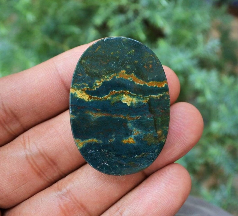 K&ouml;nnte beinhalten: Ein ovaler Edelstein mit einer dunkelblauen Grundfarbe, der goldene und braune B&auml;nder aufweist. Der polierte Stein hat ein nat&uuml;rliches, erdiges Aussehen. Der Edelstein wird in einer Hand gehalten.