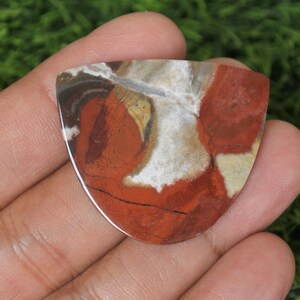 Op de afbeelding: Een rode, witte en bruine agaat cabochon met een uniek, swirlend patroon. De cabochon is traanvormig en heeft een glad, gepolijst oppervlak.