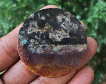 Puede incluir: Una piedra redonda y pulida con un patrón de color único. La mitad superior es oscura con motas más claras, que transicionan a una base marrón rojizo y amarilla. La superficie de la piedra tiene una apariencia natural y texturizada, con una mezcla de colores.