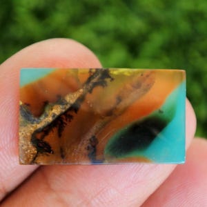 May include: A rectangular gemstone with a vibrant mix of colors. The stone features a base of turquoise, with layers of orange, brown, and black, and a touch of gold. The stone is held in a hand.
