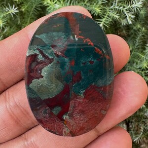May include: An oval-shaped gemstone with a polished surface. The stone features a blend of deep red, forest green, and gray hues, creating a marbled effect. The gemstone is held in a hand, showcasing its natural beauty and unique color variations.