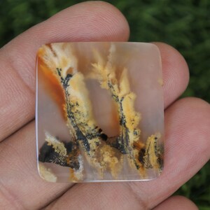 May include: Square-shaped gemstone with a unique pattern. The stone features a blend of colors, including beige, orange, yellow, and black, creating a natural, artistic design. The gemstone is held in a hand, showcasing its size and detail.