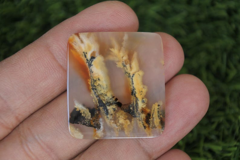 May include: Square-shaped gemstone with a unique pattern. The stone features a blend of colors, including beige, orange, yellow, and black, creating a natural, artistic design. The gemstone is held in a hand, showcasing its size and detail.