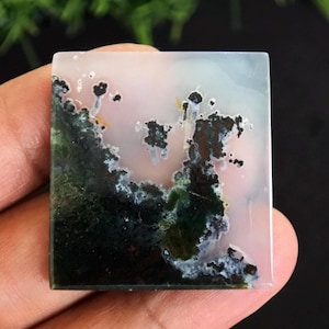Peut inclure: Une pierre précieuse carrée avec un motif unique. La pierre présente un mélange de couleurs, dont le vert, le noir, le blanc et le rose. Le motif ressemble à un paysage avec des zones sombres et moussues et des formations plus claires en forme de nuages. La pierre est tenue dans une main.