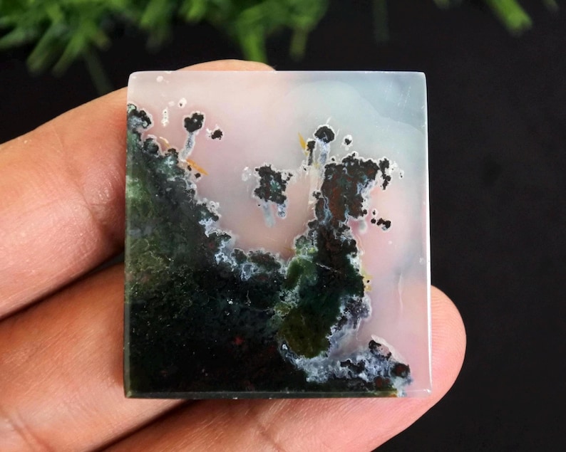 Puede incluir: Una piedra preciosa cuadrada con un patr&oacute;n &uacute;nico. La piedra presenta una mezcla de colores, incluyendo verde, negro, blanco y rosa. El dise&ntilde;o se asemeja a un paisaje con &aacute;reas oscuras y musgosas y formaciones m&aacute;s claras en forma de nube. La piedra se sostiene en una mano.