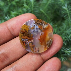 May include: A round, polished agate cabochon with a mix of white, brown, orange, and red colours. The agate has a unique, swirling pattern.