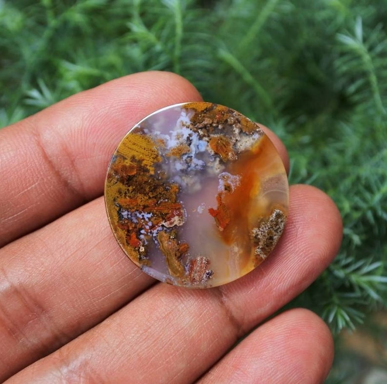 May include: A round, polished agate cabochon with a mix of white, brown, orange, and red colours. The agate has a unique, swirling pattern.