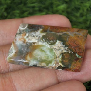 K&ouml;nnte beinhalten: Ein rechteckiger, gr&uuml;n-braun-wei&szlig;er Achat-Cabochon mit polierter Oberfl&auml;che. Der Stein hat ein einzigartiges Muster aus Wirbeln und Flecken.