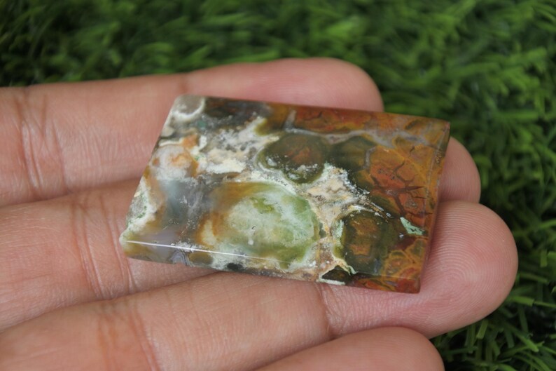 K&ouml;nnte beinhalten: Ein rechteckiger, gr&uuml;n-braun-wei&szlig;er Achat-Cabochon mit polierter Oberfl&auml;che. Der Stein hat ein einzigartiges Muster aus Wirbeln und Flecken.