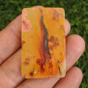 May include: A rectangular polished gemstone cabochon featuring a warm color palette of golden yellow, burnt orange, and soft pink tones. The surface displays a prominent dark brown, vertical dendritic inclusion that resembles a branching tree.