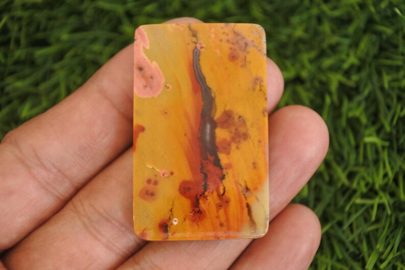 May include: A rectangular polished gemstone cabochon featuring a warm color palette of golden yellow, burnt orange, and soft pink tones. The surface displays a prominent dark brown, vertical dendritic inclusion that resembles a branching tree.