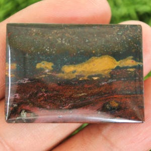 Może przedstawiać: Prostokątny kamień szlachetny o wypolerowanej powierzchni. Kamień ma wzór przypominający krajobraz z kolorami czerwonym, żółtym, niebieskim i brązowym. Kamień szlachetny jest trzymany w dłoni na rozmytym zielonym tle.
