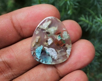 May include: A cabochon cut gemstone with a white, gray, and brown base with green and red inclusions. The stone is oval shaped and has a smooth, polished surface.