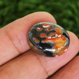 Może przedstawiać: Wypolerowany, owalny kamień szlachetny z mieszanką kolor&oacute;w. Kamień ma błyszczącą powierzchnię z mieszanką czarnych, pomarańczowych i białych odcieni. Kamień szlachetny jest trzymany w dłoni, prezentując swoją gładką fakturę i unikalne wzory kolorystyczne.