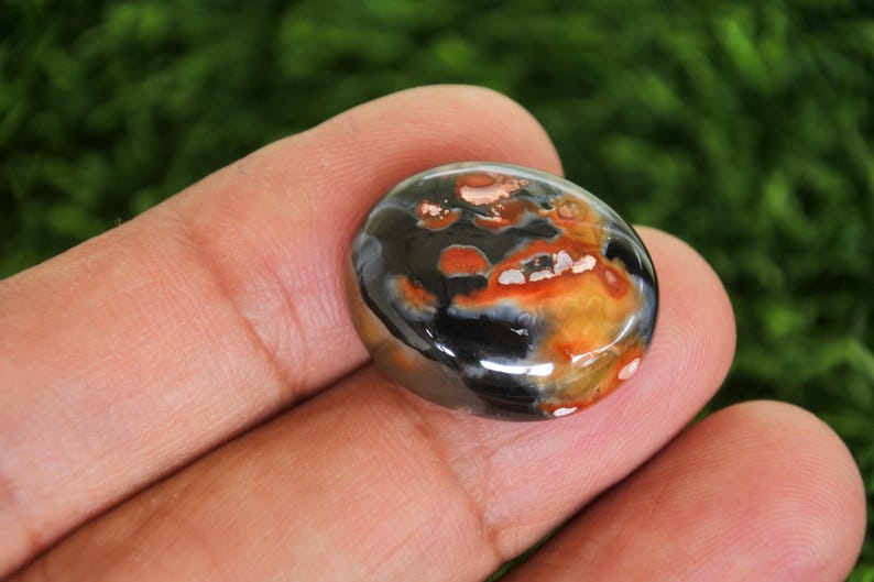 Może przedstawiać: Wypolerowany, owalny kamień szlachetny z mieszanką kolor&oacute;w. Kamień ma błyszczącą powierzchnię z mieszanką czarnych, pomarańczowych i białych odcieni. Kamień szlachetny jest trzymany w dłoni, prezentując swoją gładką fakturę i unikalne wzory kolorystyczne.