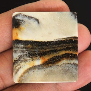 May include: A square, polished stone with a distinctive pattern. The stone displays layers of cream, black, orange, and grey, forming a landscape-like design. The edges are smooth and rounded, held in a hand.