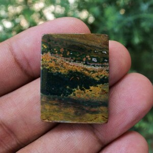 Pode incluir: Uma pedra preciosa retangular com um padr&atilde;o natural de tons verde escuro, castanho e amarelo. A pedra tem uma superf&iacute;cie polida e uma mistura de cores e texturas, criando uma apar&ecirc;ncia &uacute;nica. A pedra preciosa &eacute; segurada em uma m&atilde;o.