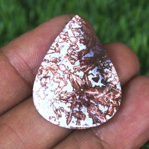 May include: A teardrop-shaped gemstone with a unique pattern of white, brown, and blue hues. The stone's surface displays a complex, natural design, resembling a landscape or abstract art. The gemstone is held in a hand, showcasing its size and shape.
