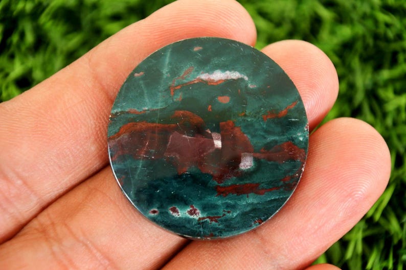 Peut inclure: Une pierre pr&eacute;cieuse ronde et polie, m&eacute;langeant des couleurs vert fonc&eacute; et marron. La pierre pr&eacute;sente un aspect mouchet&eacute; avec des motifs tourbillonnants. Elle est tenue dans une main sur un fond vert flou.