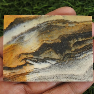 May include: A rectangular slab of polished stone featuring a natural swirling pattern of earthy tones including deep brown, ochre yellow, cream, and charcoal grey. The surface displays a smooth, matte finish with visible mineral grain textures.