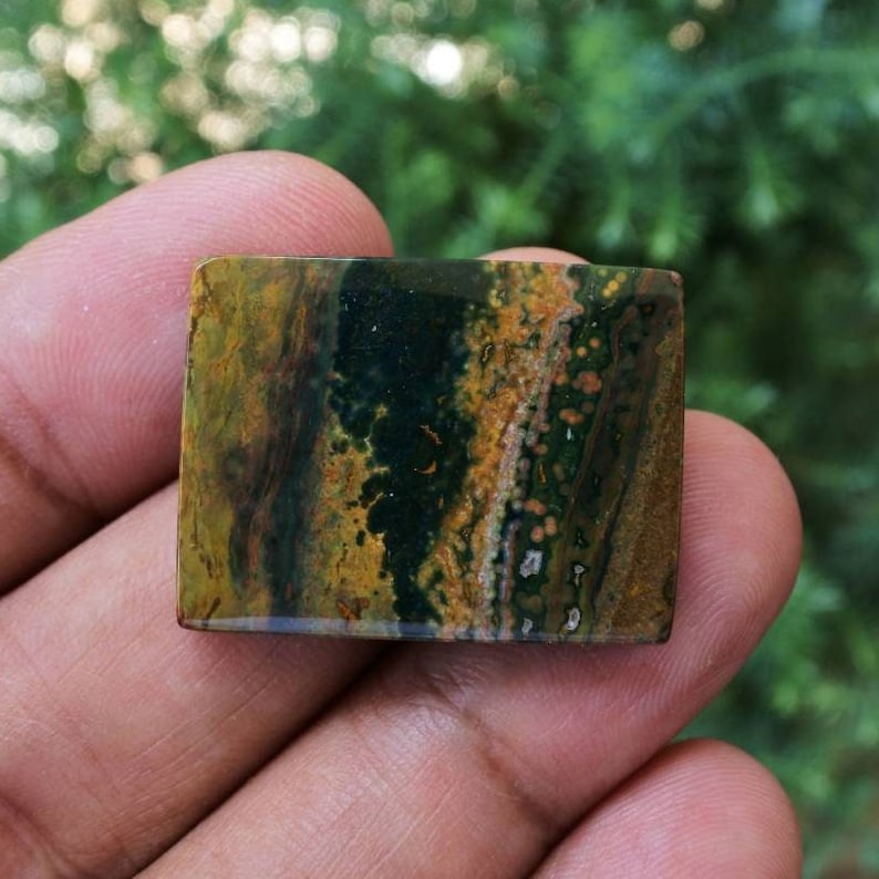 Pode incluir: Uma pedra preciosa retangular com uma superf&iacute;cie polida. A pedra apresenta uma mistura de cores, incluindo verde escuro, castanho e amarelo, com um padr&atilde;o salpicado. A pedra preciosa &eacute; segurada em uma m&atilde;o, com um fundo verde desfocado.