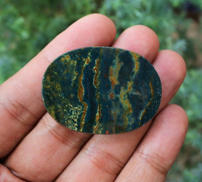 K&ouml;nnte beinhalten: Ein ovales Schmuckst&uuml;ck mit dunkelgr&uuml;ner Grundfarbe und komplizierten Mustern aus goldenen und braunen Adern. Der polierte Stein wird in einer Hand gehalten und zeigt seine nat&uuml;rliche Sch&ouml;nheit. Ca. 5 cm lang.