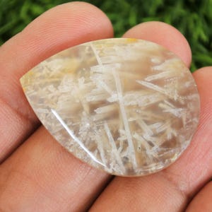 Peut inclure: Une pierre pr&eacute;cieuse en forme de goutte d'eau avec une base translucide brun clair. Des inclusions blanches et plumeuses cr&eacute;ent un motif unique sur la surface. La pierre est tenue dans une main, mettant en valeur sa finition polie.