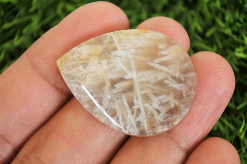 Peut inclure: Une pierre pr&eacute;cieuse en forme de goutte d'eau avec une base translucide brun clair. Des inclusions blanches et plumeuses cr&eacute;ent un motif unique sur la surface. La pierre est tenue dans une main, mettant en valeur sa finition polie.