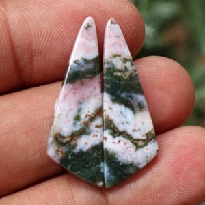 May include: Two polished, triangular gemstones with a mix of colours. The stones feature a base of white and pink, with green and brown veining. The gemstones are held in a hand.