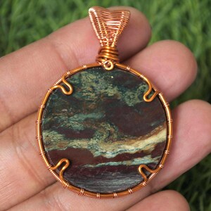 Puede incluir: Colgante redondo con marco de alambre de cobre. El colgante presenta una piedra con tonos verdes, marrones y rojos. El alambre está intrincadamente tejido alrededor de la piedra y forma un lazo en la parte superior para colgar. Joyería.
