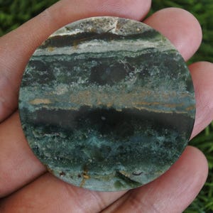 Op de afbeelding: Een ronde, gepolijste steen met een gelaagd patroon van donkergroene, blauwgroene, bruine en cr&egrave;mekleuren. De steen wordt in een hand gehouden, waarbij de natuurlijke texturen worden benadrukt. Het oppervlak van de steen is glad en gepolijst, ongeveer 7 cm in diameter.