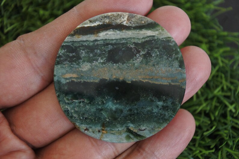 Op de afbeelding: Een ronde, gepolijste steen met een gelaagd patroon van donkergroene, blauwgroene, bruine en cr&egrave;mekleuren. De steen wordt in een hand gehouden, waarbij de natuurlijke texturen worden benadrukt. Het oppervlak van de steen is glad en gepolijst, ongeveer 7 cm in diameter.