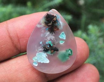 May include: A teardrop-shaped gemstone with a translucent pink base. The stone features inclusions of black, white, and turquoise-colored minerals, creating a unique pattern. The gemstone is held between fingers, with a blurred green background.