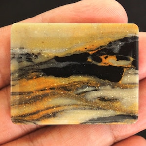 May include: A rectangular stone with a polished surface. The stone features a landscape-like pattern with layers of black, gold, and tan colours. The stone is held in a hand.
