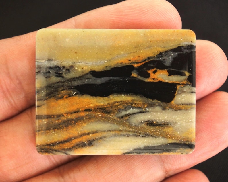 May include: A rectangular stone with a polished surface. The stone features a landscape-like pattern with layers of black, gold, and tan colors. The stone is held in a hand.