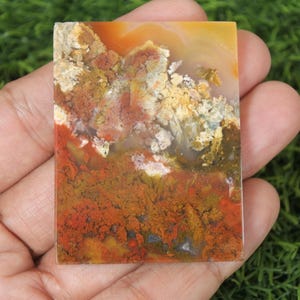 K&ouml;nnte beinhalten: Ein polierter Achat-Cabochon mit einem farbenfrohen Muster aus Rot, Orange, Gelb und Braun. Der Stein hat ein nat&uuml;rliches, organisches Aussehen.