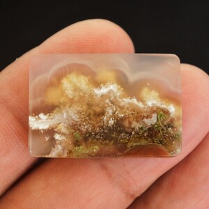 May include: A rectangular gemstone with a translucent appearance and a landscape design. The stone displays a blend of colours, including white, brown, yellow, and green, reminiscent of a cloudy sky over a landscape. The stone is held in a hand.