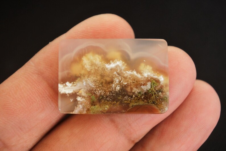 May include: A rectangular gemstone with a translucent appearance and a landscape design. The stone displays a blend of colours, including white, brown, yellow, and green, reminiscent of a cloudy sky over a landscape. The stone is held in a hand.