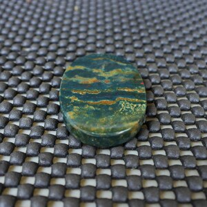 K&ouml;nnte beinhalten: Ein oval geformter Edelstein mit polierter Oberfl&auml;che. Der Stein weist eine Mischung aus dunkelgr&uuml;nen, braunen und rot-orangen Farbt&ouml;nen auf, die ein nat&uuml;rliches Muster bilden. Der Edelstein wird wahrscheinlich f&uuml;r Schmuck oder Dekorationszwecke verwendet.