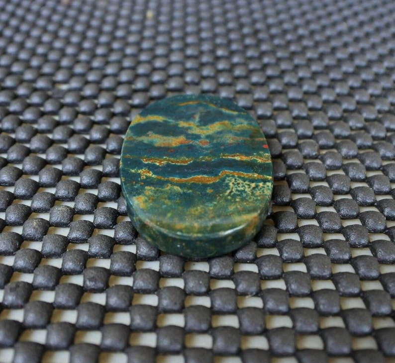 K&ouml;nnte beinhalten: Ein oval geformter Edelstein mit polierter Oberfl&auml;che. Der Stein weist eine Mischung aus dunkelgr&uuml;nen, braunen und rot-orangen Farbt&ouml;nen auf, die ein nat&uuml;rliches Muster bilden. Der Edelstein wird wahrscheinlich f&uuml;r Schmuck oder Dekorationszwecke verwendet.