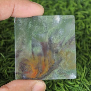 Peut inclure: Une pierre carr&eacute;e polie avec un motif tourbillonnant de teintes vertes, jaunes, oranges et violettes. La pierre est tenue entre deux doigts, mettant en valeur ses couleurs et textures naturelles. La surface de la pierre est lisse et r&eacute;fl&eacute;chissante.