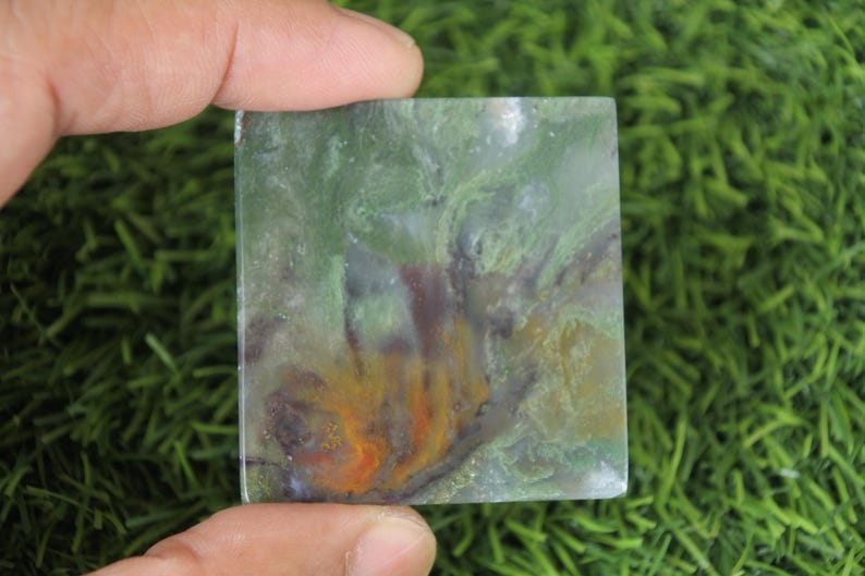 Peut inclure: Une pierre carr&eacute;e polie avec un motif tourbillonnant de teintes vertes, jaunes, oranges et violettes. La pierre est tenue entre deux doigts, mettant en valeur ses couleurs et textures naturelles. La surface de la pierre est lisse et r&eacute;fl&eacute;chissante.